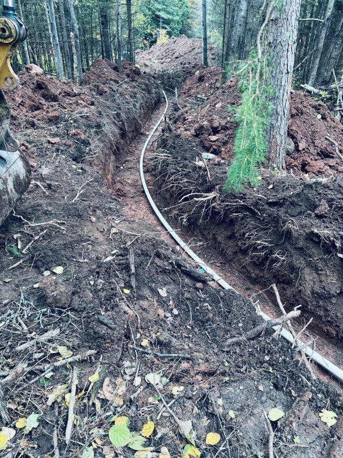 A white pipe curves in a dirt trench through a forest. Construction in progress.
