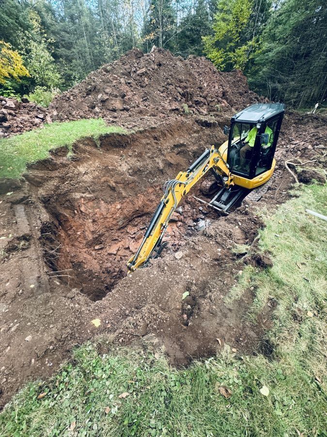 Yellow excavator digging a large rectangular hole in a grassy area. A pile of dirt sits in the background.
