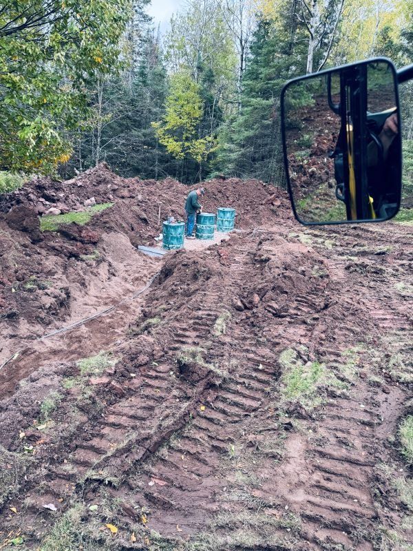 Person near blue barrels in a dirt excavation, with tracks from heavy machinery. Forest in background.