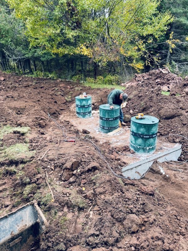 A person installing a septic system, surrounded by dirt and green tanks, in a wooded area.