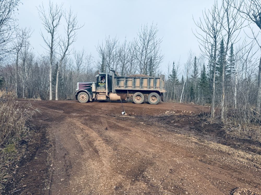 Dump truck on a dirt road in a wooded area, hauling dirt, overcast sky.