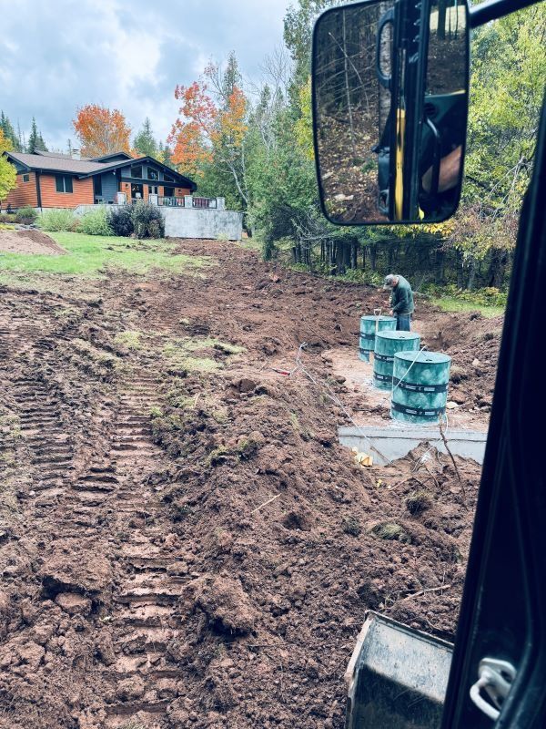 A construction site, earthmoving equipment, with a house in the background and a worker near precast concrete.