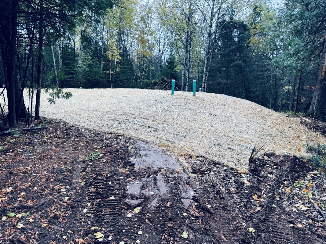 An earthen mound with straw, blue pipes, surrounded by trees and a muddy foreground.