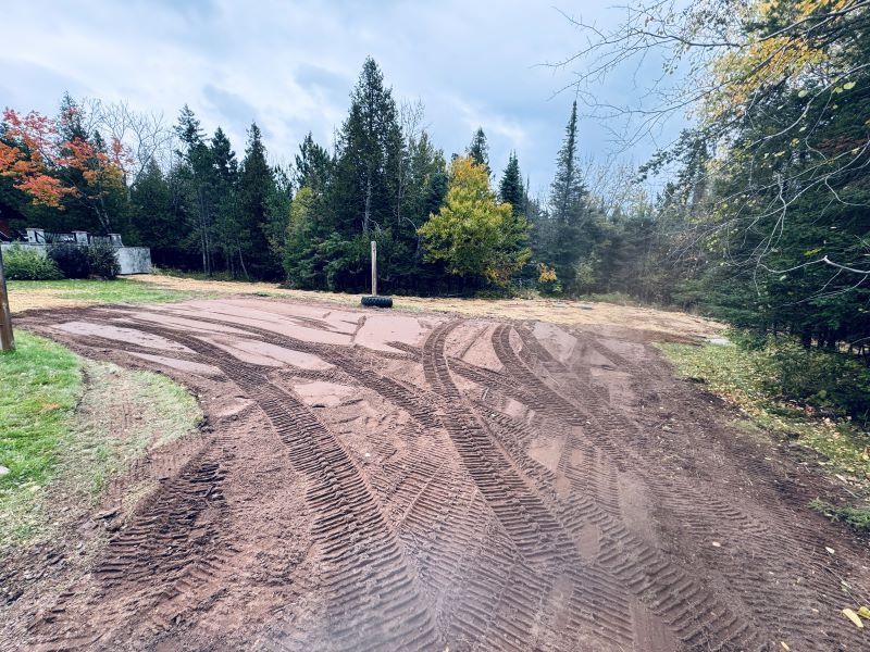 Muddy driveway with tire tracks, leading to trees and a cloudy sky.