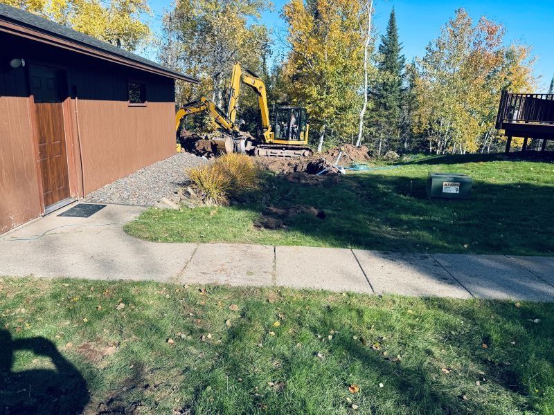 Excavator digging near a brown building and a sidewalk. Autumn leaves and green grass.