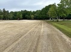 Gravel road leading through a large, empty gravel lot, bordered by green grass and trees under a cloudy sky.