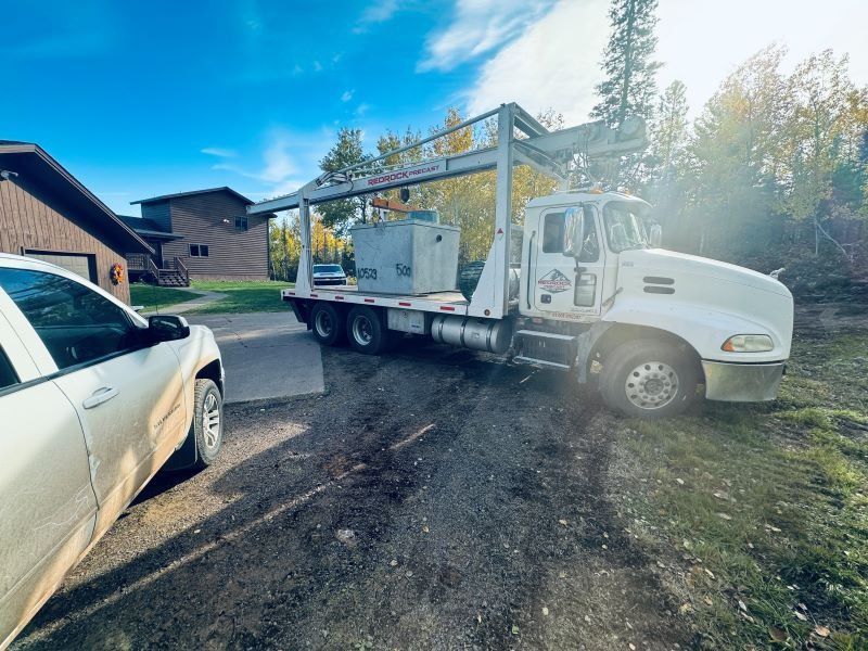 A white truck with a large concrete container on its flatbed in front of a house.
