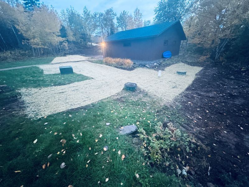 Gravel pathways and grass yard lead to a small brown shed, with trees in the background.