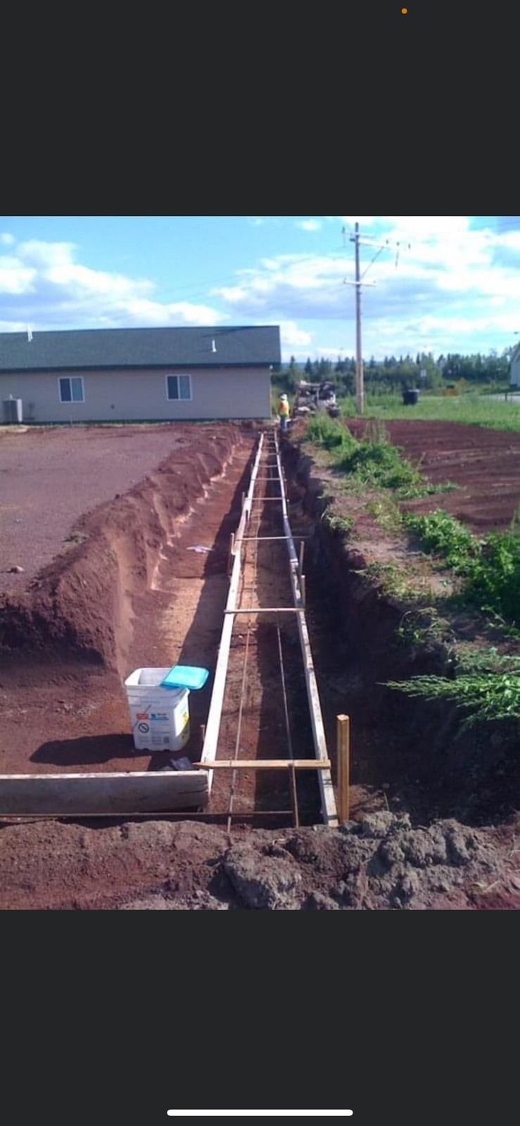 Construction site: Long trench alongside a building, with wooden frames and exposed soil, under a blue sky.