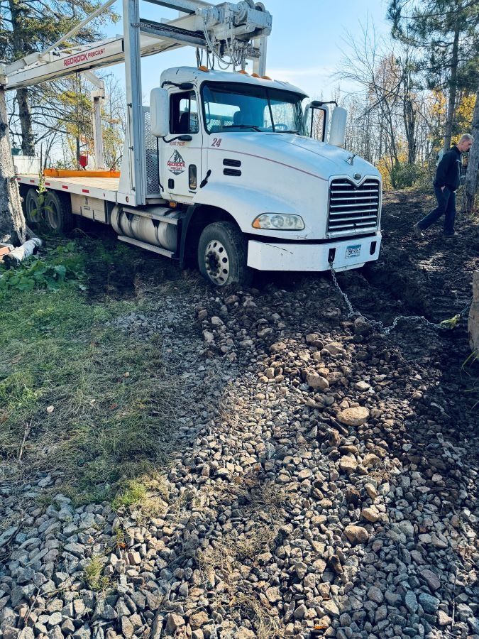 White truck stuck in mud, with a boom, in a wooded area; a person walks nearby.