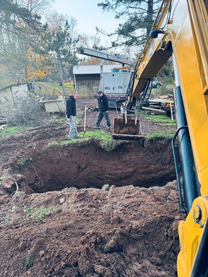 A backhoe digs a large hole, two men watch in a yard, surrounded by dirt, trees, and buildings.