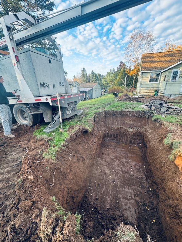 Large pit dug in yard; truck with lift arm nearby, man standing.