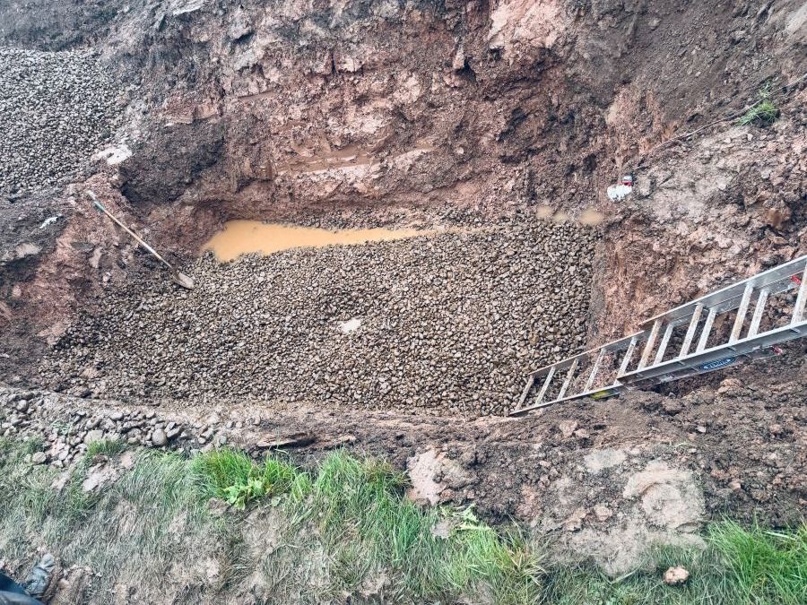 Excavated bank filled with rocks, muddy water at top, ladder at right, green grass below.