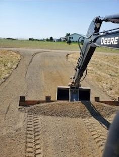 A John Deere backhoe grading a gravel driveway with tire tracks in a rural setting.