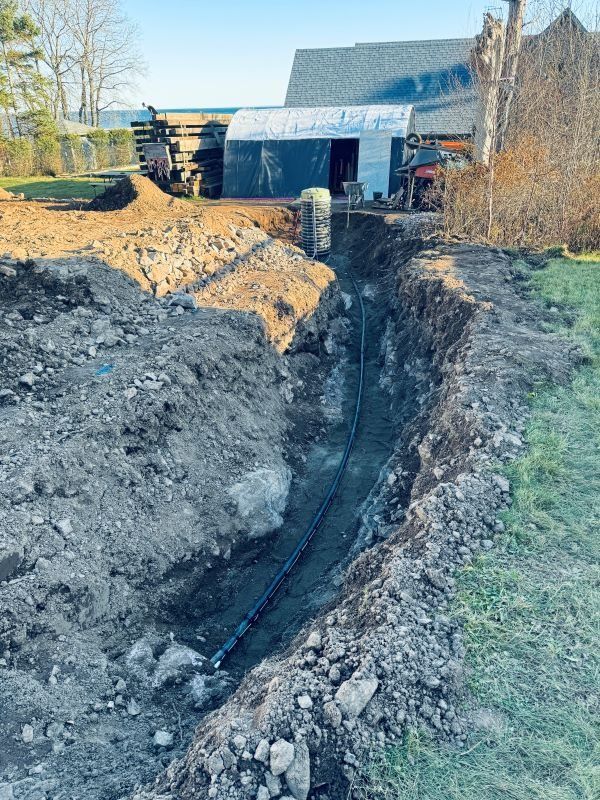 A trench dug in a yard, containing a black pipe, leading toward a building.