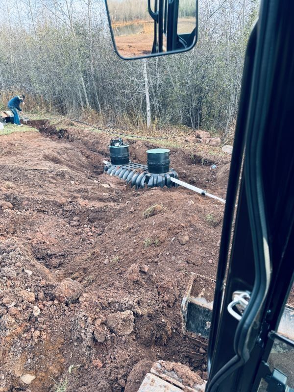 Excavator view of a septic system installation; trenches, tanks, piping in a brown dirt field, workers in the background.
