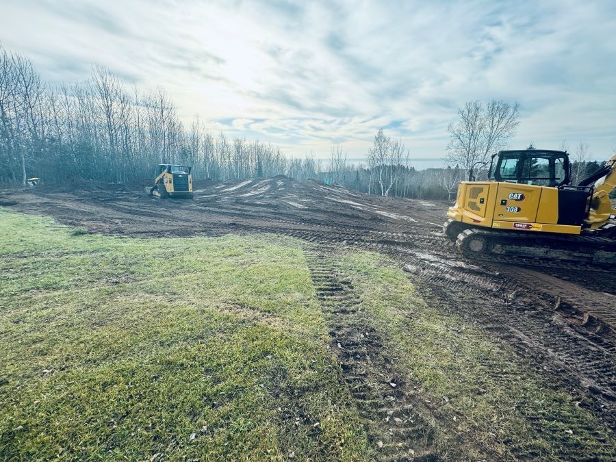 Construction site with excavator and skid steer on muddy ground near trees.