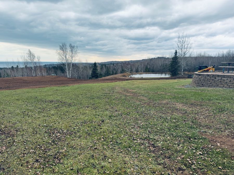 Grassy field with a pond and trees under a cloudy sky. Lake in the distance.