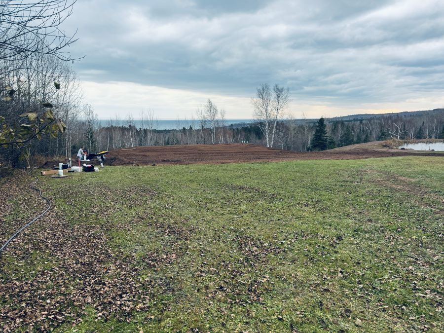 A brown field with sparse grass and view of water, trees, and cloudy sky.