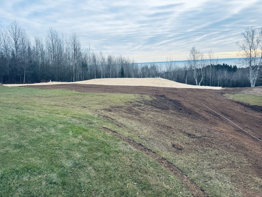 Grassy hillside with bare trees and an expanse of dirt, possibly for construction, under a cloudy sky.
