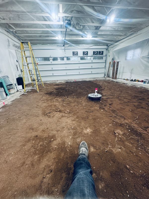 A person's legs and feet in a garage; robot vacuum on the dirt floor, ladder at left.