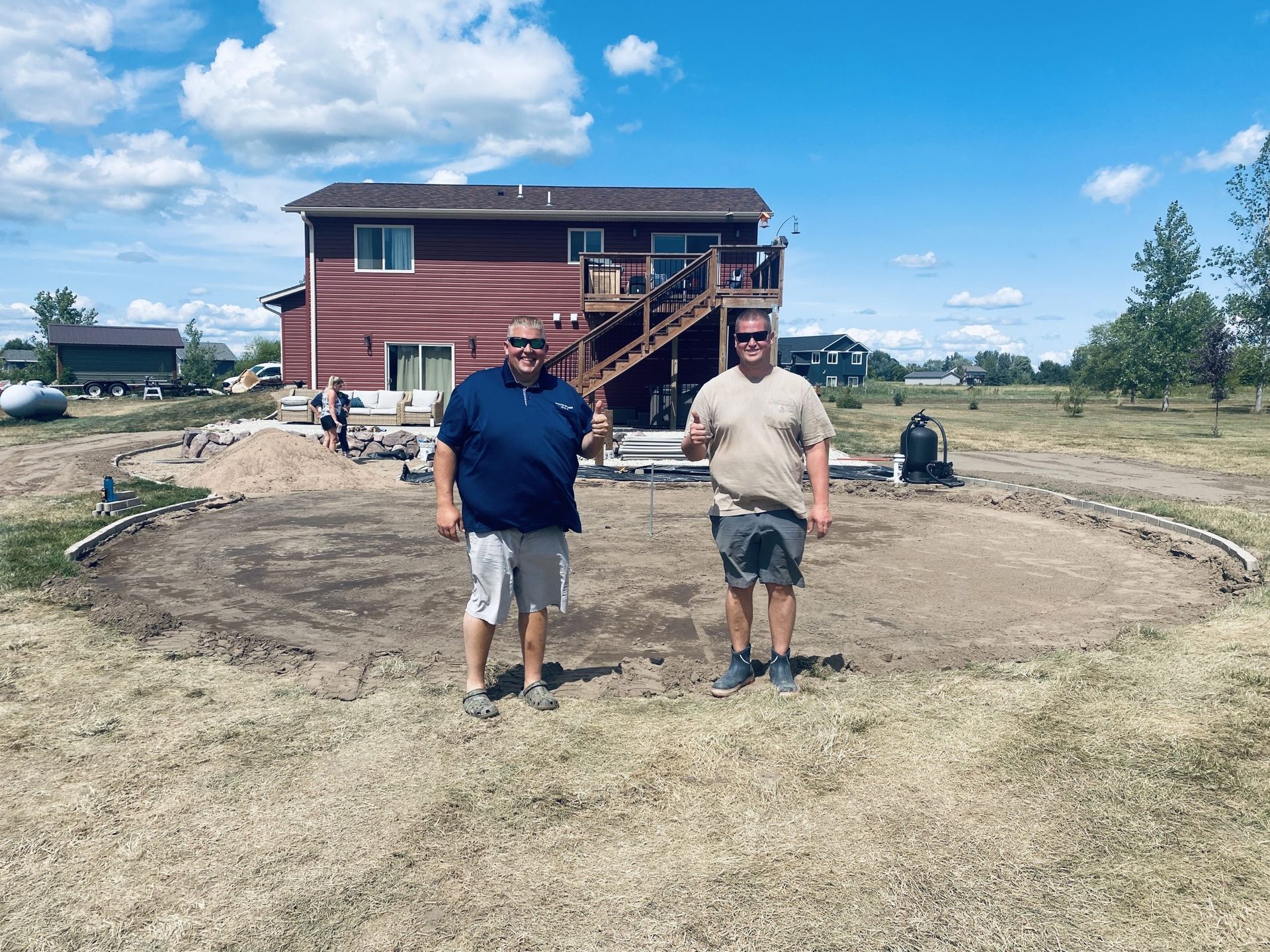 Two men stand in front of a house, smiling. A circular area of dirt suggests a future pool. Outdoors, sunny day.