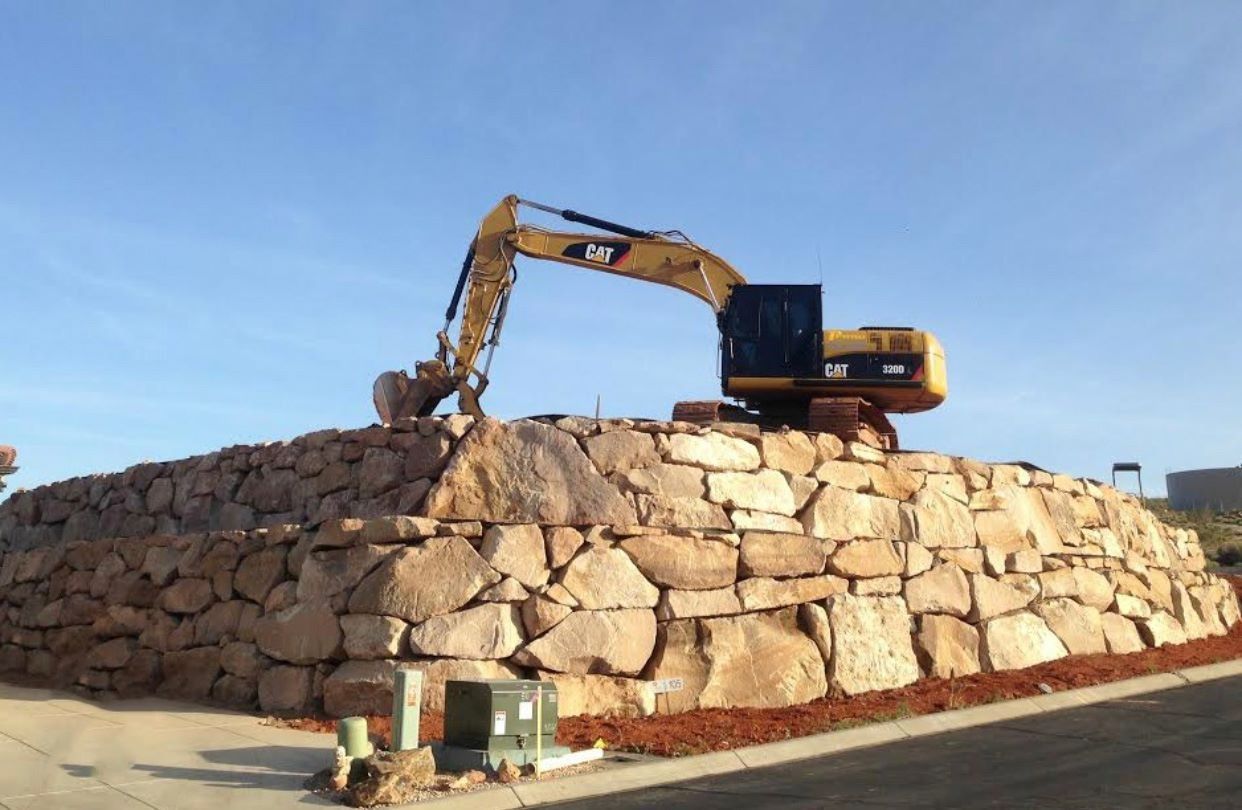 Yellow excavator on a large stone wall, under a blue sky.