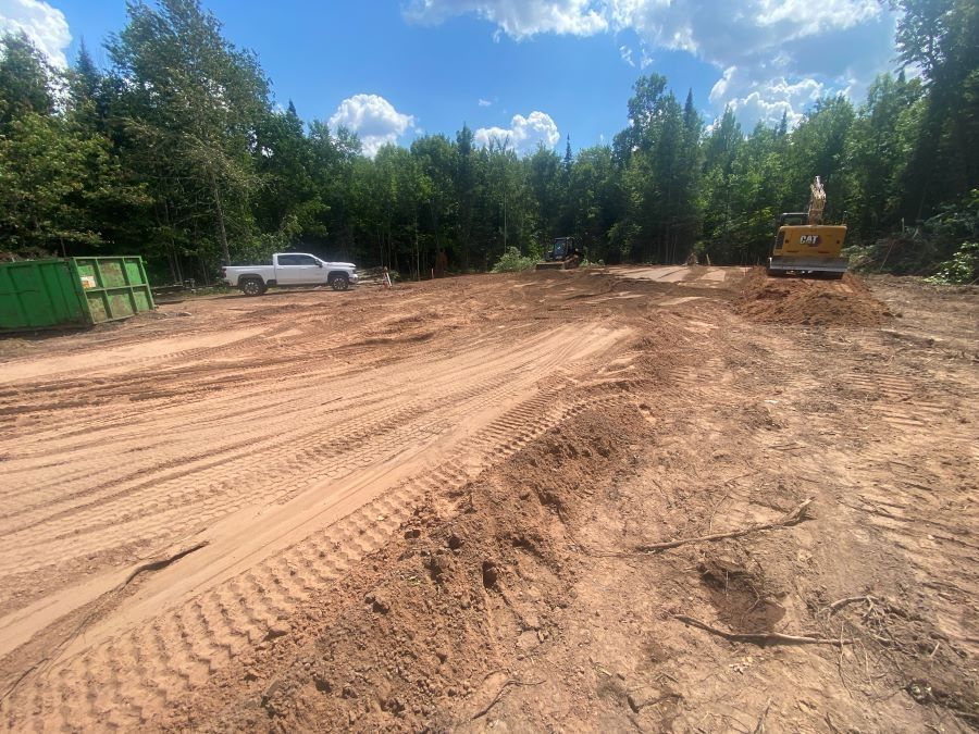 Construction site with a truck, excavator, and debris containers against a backdrop of trees.