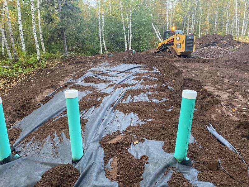 A skid steer on a construction site, covering landscape fabric with dirt and green pipes.