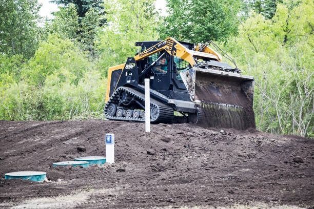 A compact track loader pushing dirt on a construction site; surrounded by trees.