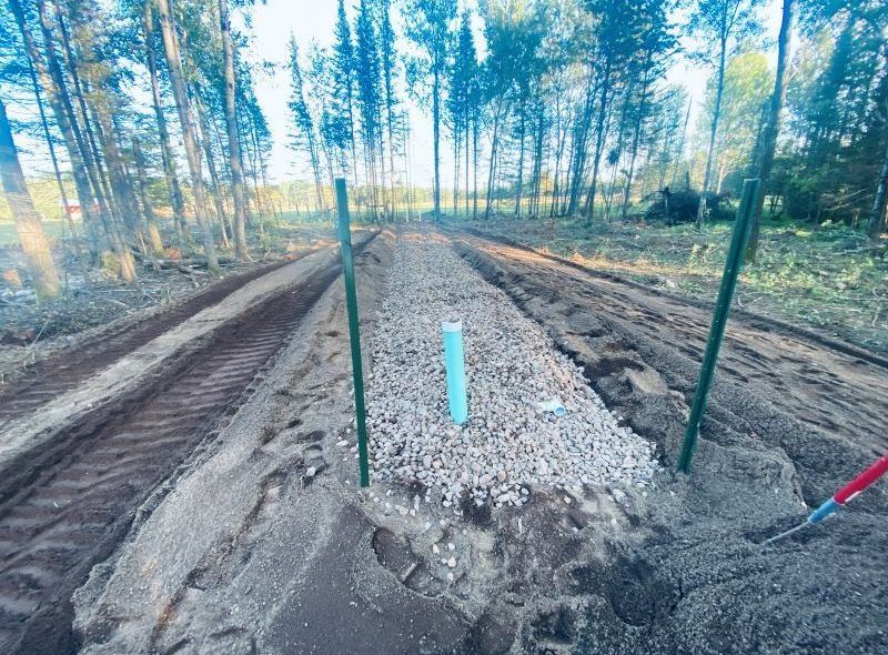 A dirt road with gravel lined trenches, green posts, and a blue pipe in a rural setting.