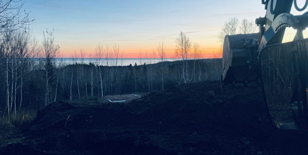 An excavator digging earth at dusk, with a colorful sunset over water and forest in the background.