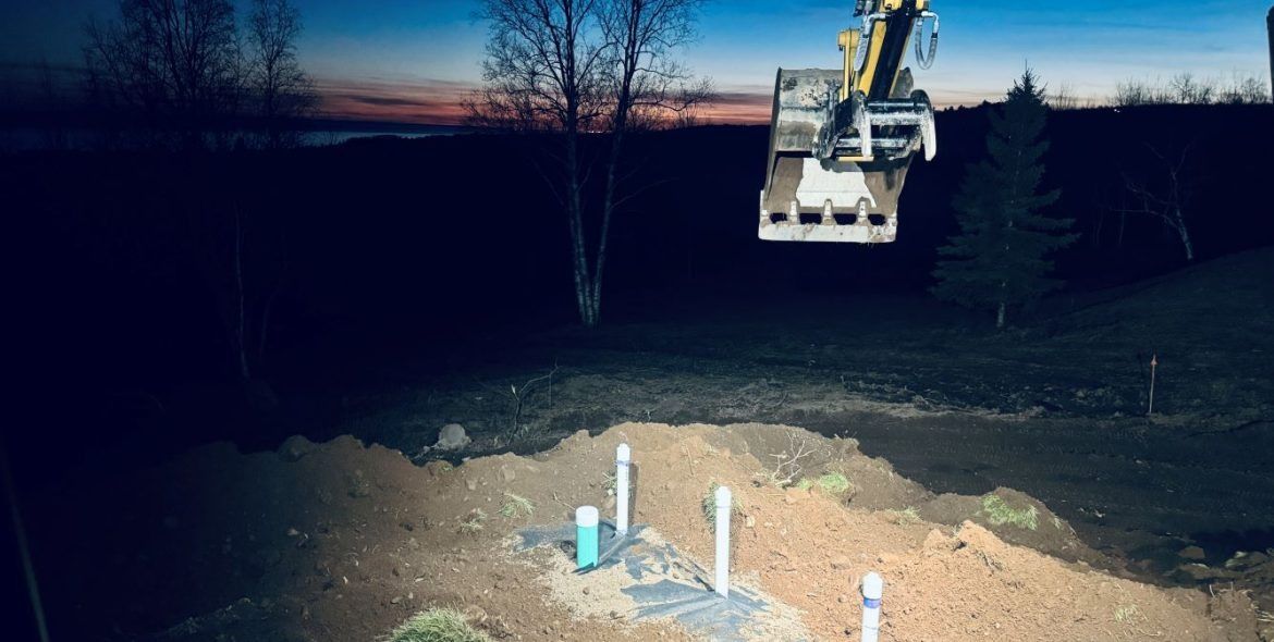 Backhoe working at dusk over excavated area with PVC pipes.
