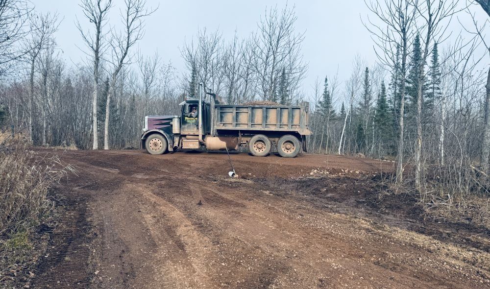 A dump truck on a dirt road in a wooded area. Gray sky, brown truck.