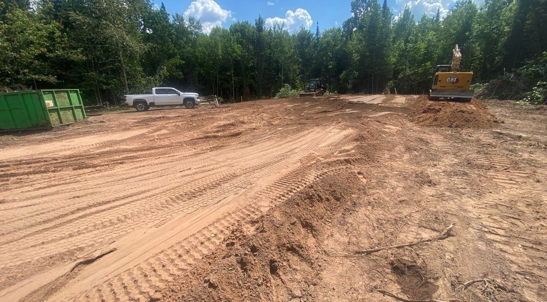 Cleared construction site with a truck, excavator, and dumpster in front of a tree line under a blue sky.