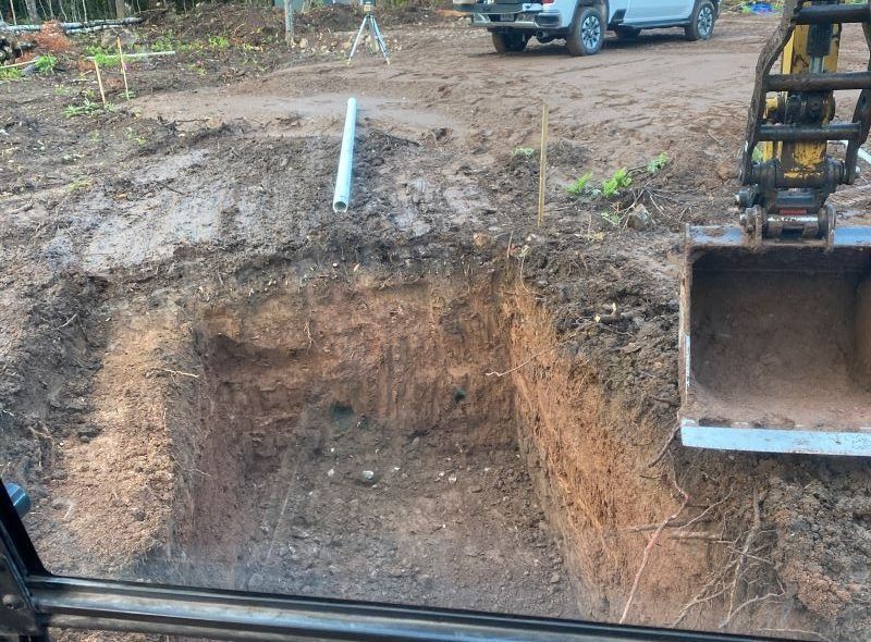 Excavation pit, revealing soil layers. Backhoe bucket to the right. White truck, survey equipment in the background.
