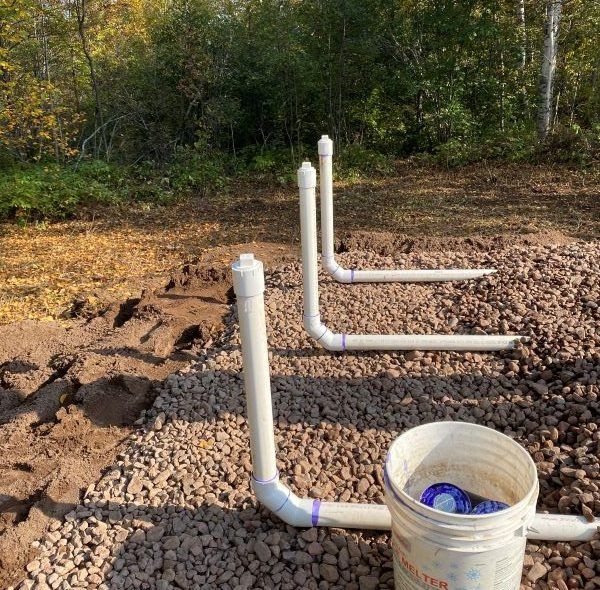 PVC pipes jut from gravel ground, next to a bucket. Forest in background.