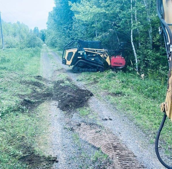 A small yellow forestry machine on a gravel road beside trees, with dirt and tire tracks visible.