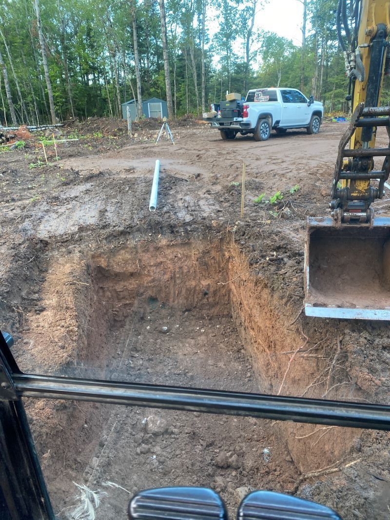 Excavator digging in dirt; truck, trees, and structure in the background on a construction site.