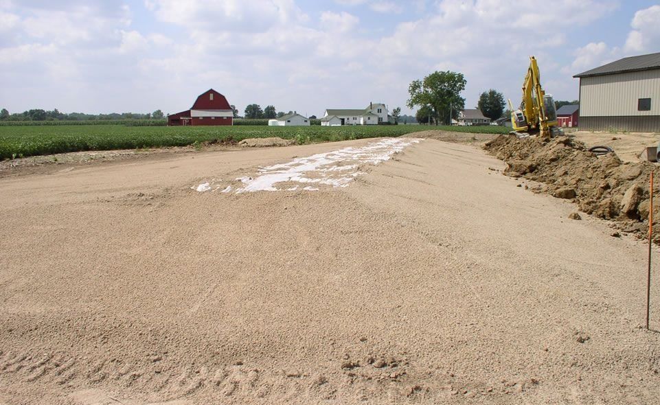 Construction site with gravel base, excavator, and rural buildings in the background.