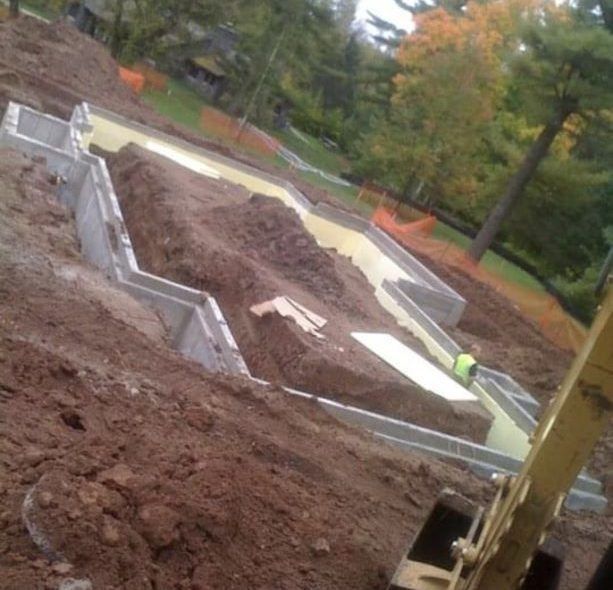 Foundation excavation for a building; dirt, concrete, orange safety netting, excavator, and a worker.