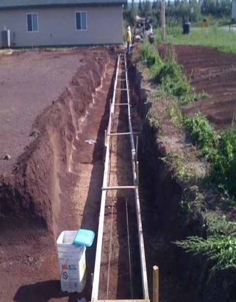 A long, deep trench with concrete forms is ready for pouring. Workers nearby, near a building and grass.