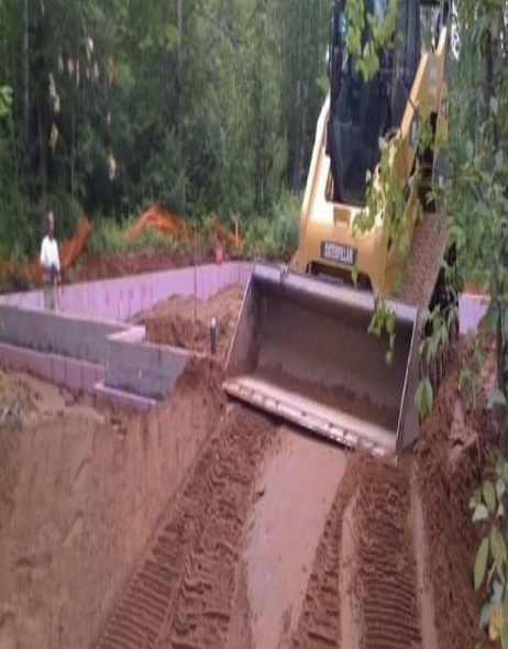A small yellow construction vehicle backfilling a foundation pit with dirt, forest background.