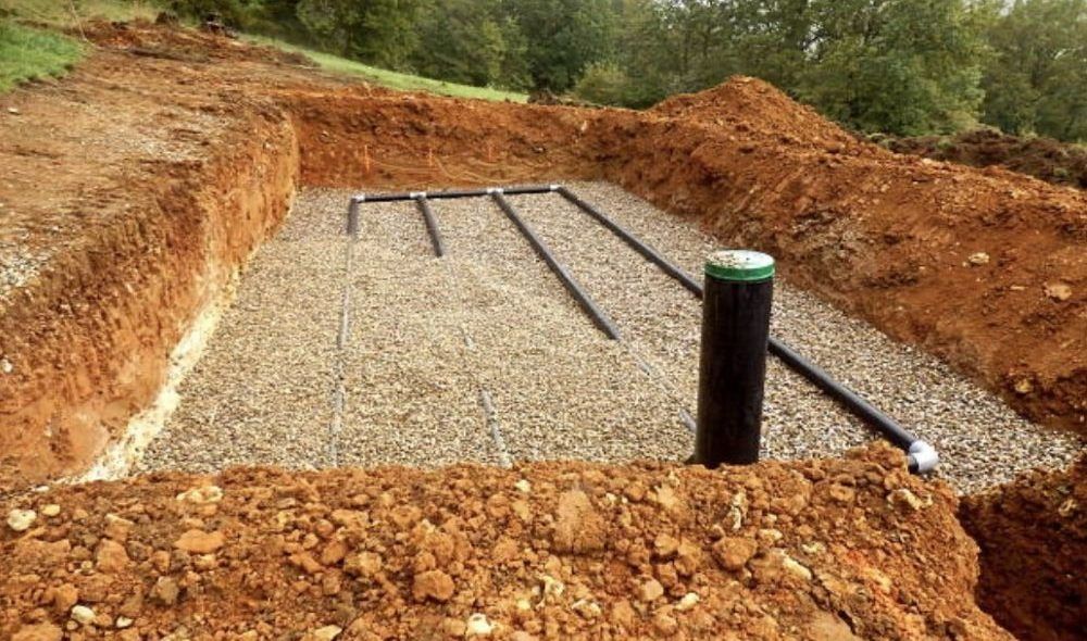 Construction of a septic system. A gravel-filled trench contains black pipes, and a black pipe extends upward.