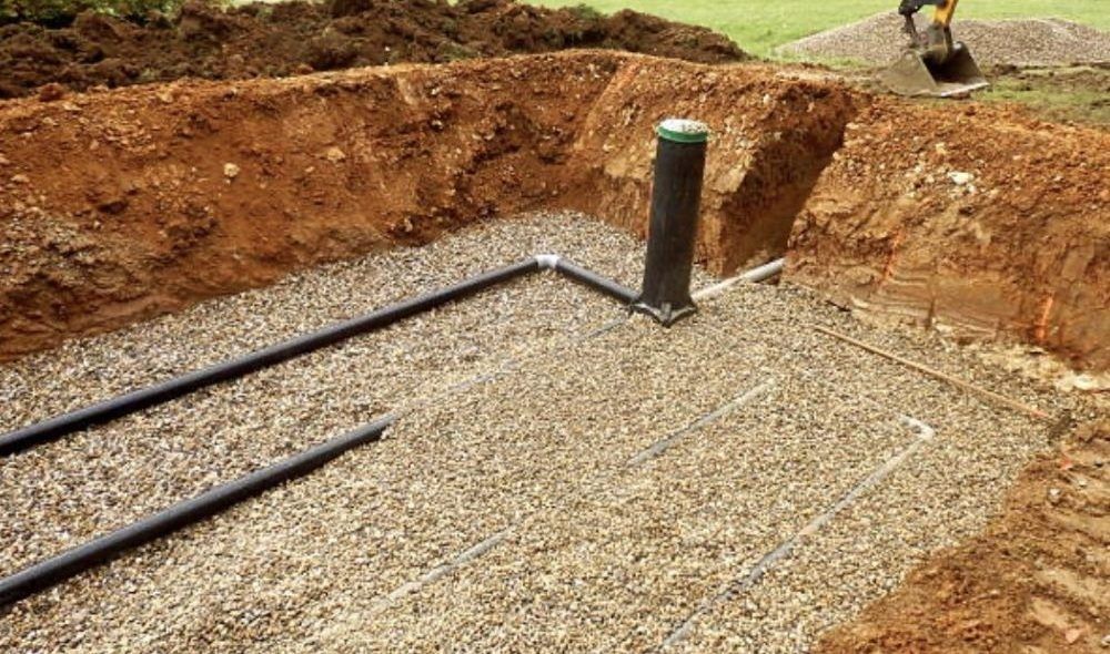 Gravel-filled trench with black pipes, and a vertical black pipe. Dirt and a backhoe in the background.