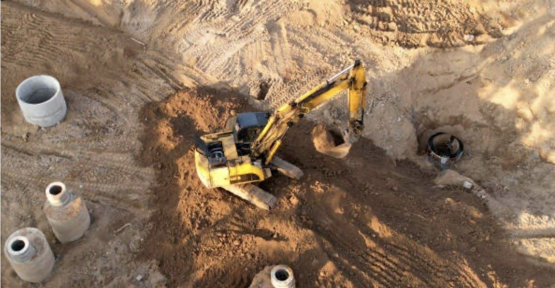 Yellow excavator digging in a sandy construction site, surrounded by concrete cylinders.