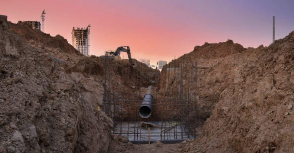 Construction site, trench with large pipe, excavator at dusk, dirt walls, orange and purple sky.
