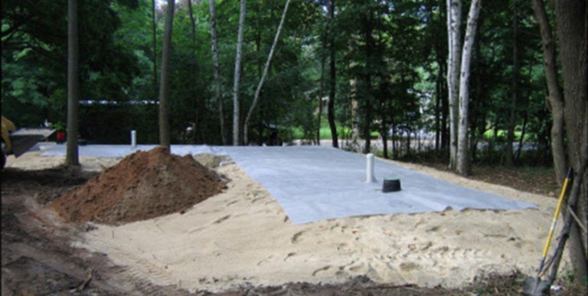 Construction site with a sand base, dirt pile, and plastic sheeting, surrounded by trees.