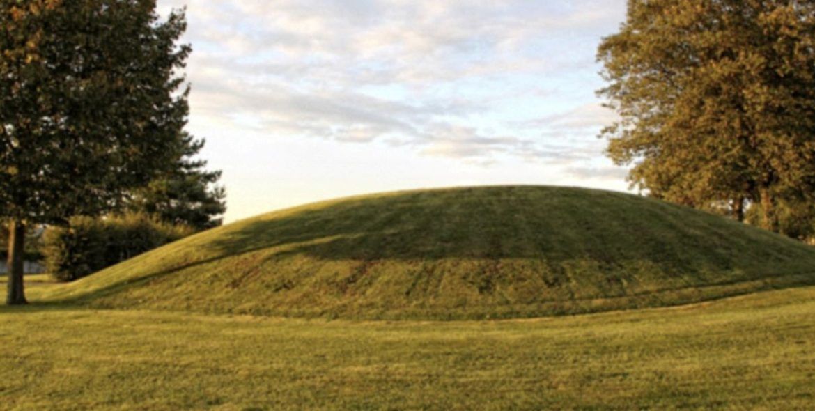 A grassy mound in a park, with trees on either side. Cloudy sky overhead.