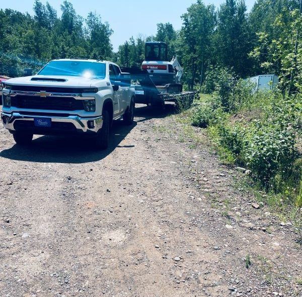 White truck towing an excavator on a trailer on a dirt road; trees and blue sky in background.